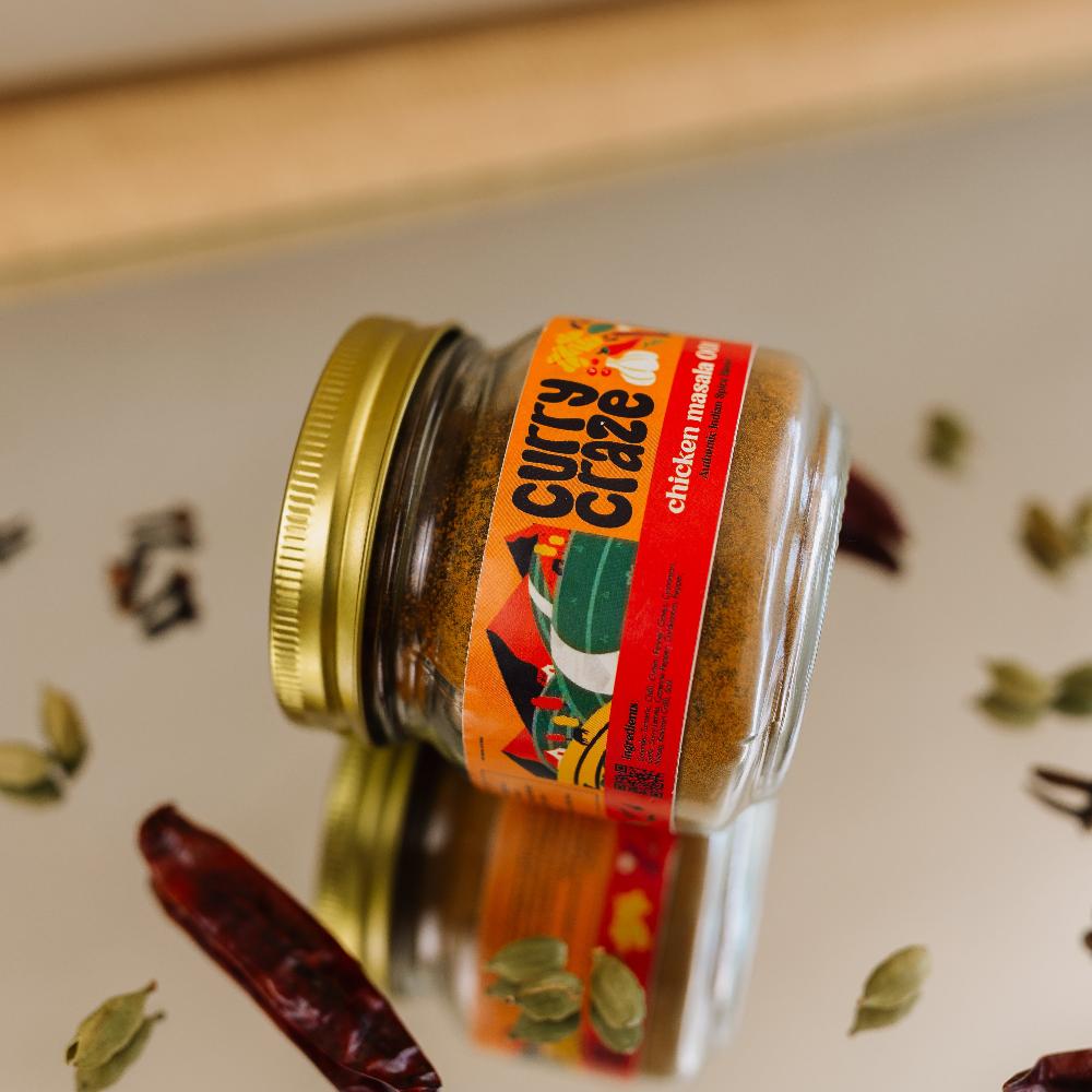 Curry Crazy chicken masala spice jar lying on its side on a reflective surface, surrounded by scattered spices like cardamom and dried chilies, enhancing the product's rich flavor profile.